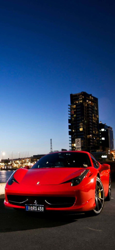 Glossy red Ferrari 458 parked by calm waterfront with cityscape and evening lights at dusk wallpaper