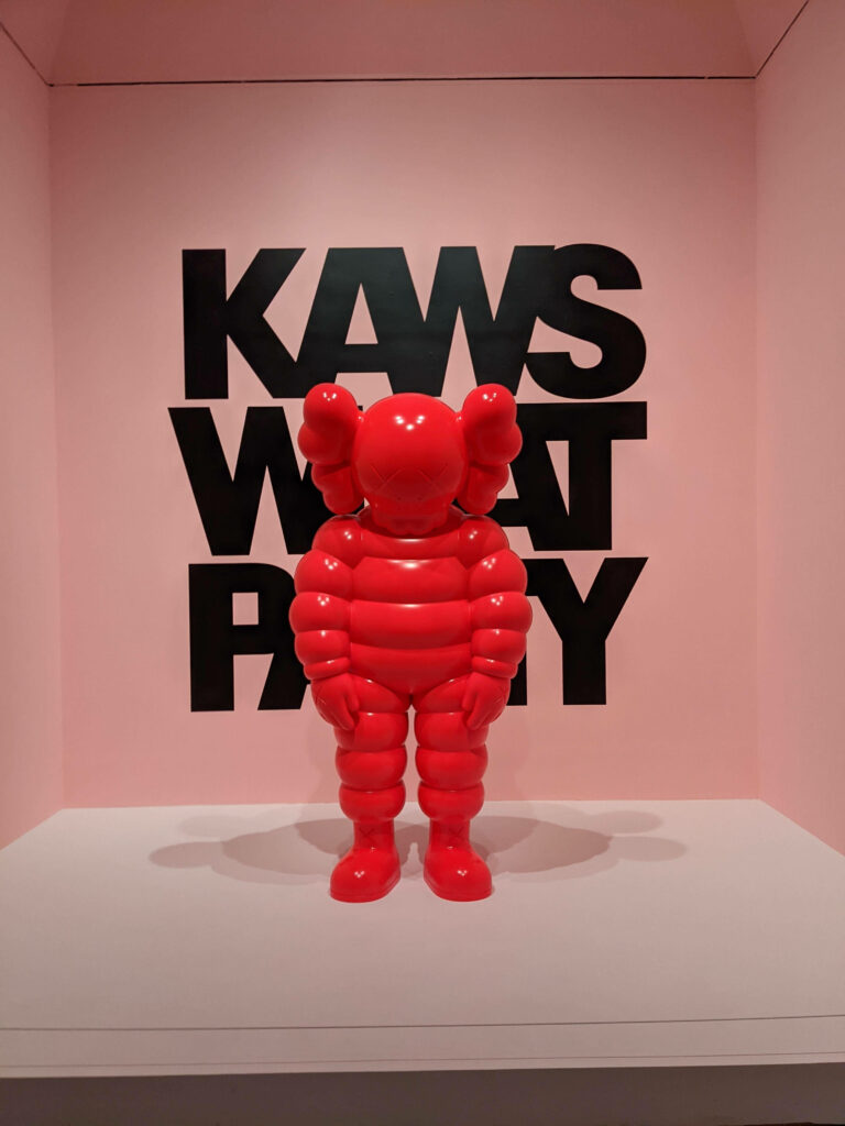 Glossy red KAWS What Party figure on soft pink background with bold black KAWS text wallpaper