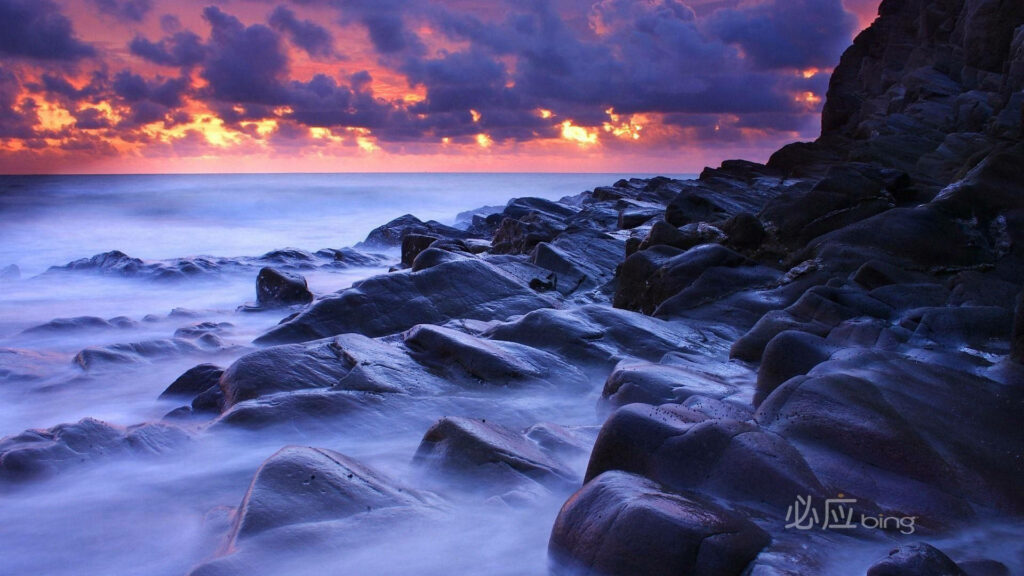 Long-exposure coastal scene with smooth rocks and vibrant orange purple sunset waves wallpaper background