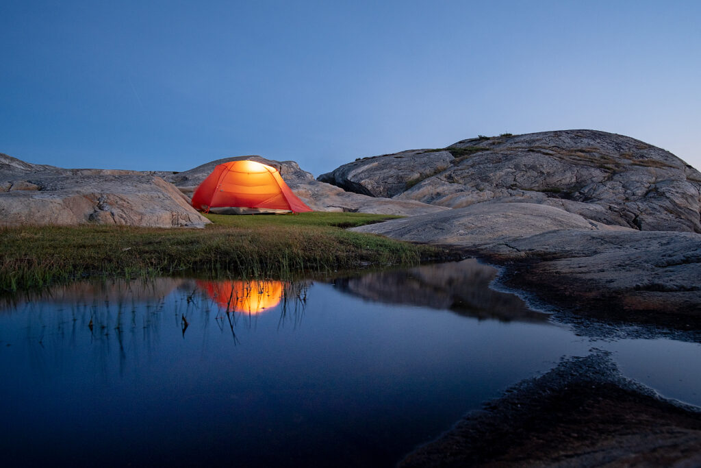 Glowing orange tent by calm pond among rugged stones at serene evening outdoor camping scene Wallpaper
