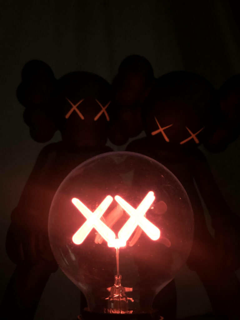 Glowing red light bulb with XX design in front of shadowy KAWS figures moody artistic wallpaper