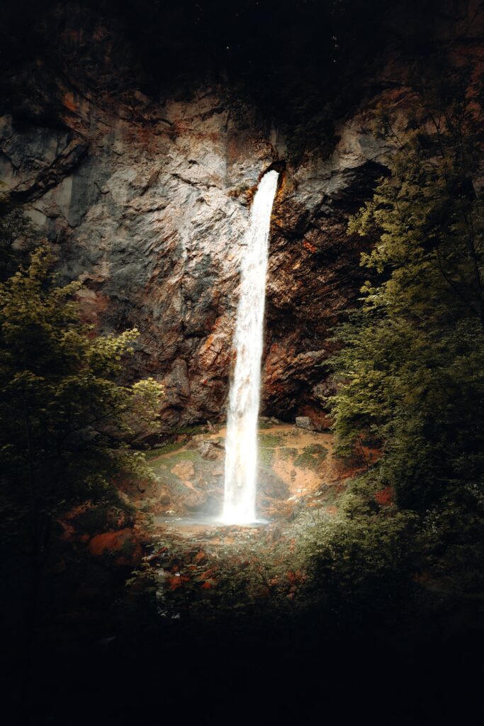 Waterfall cascading down dark rocky cliffs surrounded by lush green trees vibrant nature wallpaper