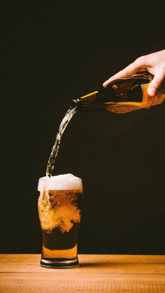 Golden ale beer pouring into glass with frothy foam on rustic wooden table wallpaper background