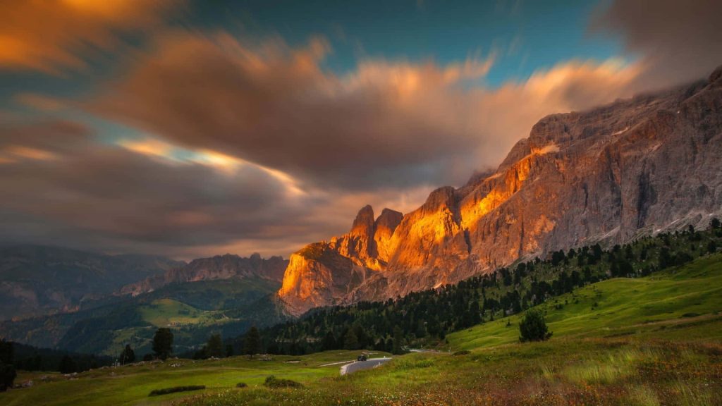 Golden sunset alpenglow on jagged Dolomite peaks with alpine meadows and winding road Wallpaper