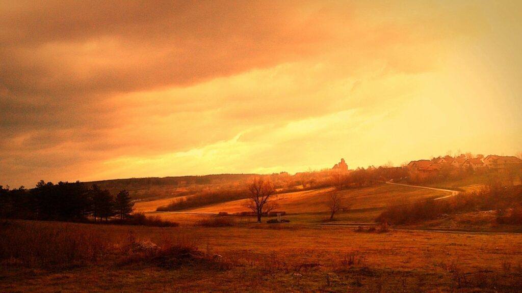 Sunlit countryside field with autumn and summer hues rolling hills trees and distant village Wallpaper