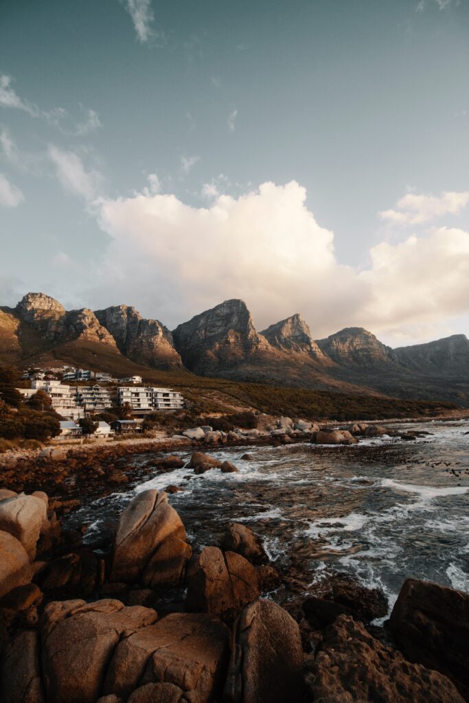 Dramatic coastline with rugged rocks crashing waves majestic mountains modern buildings golden sunlight wallpaper
