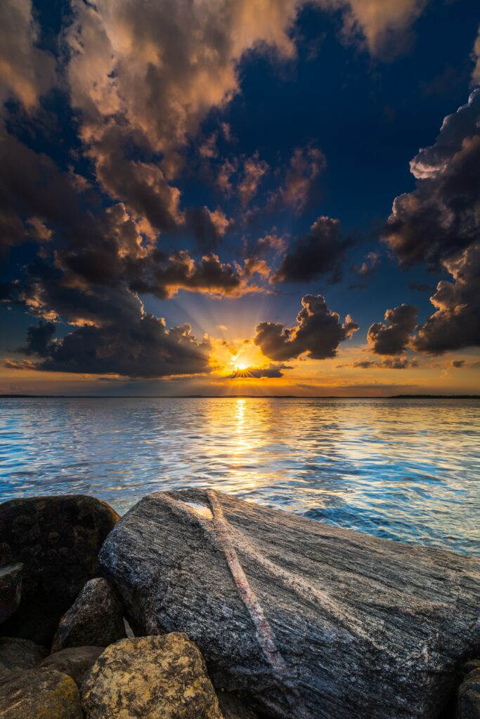 Lakeside dawn landscape with golden sunlight through clouds reflecting on calm water and rocks Wallpaper