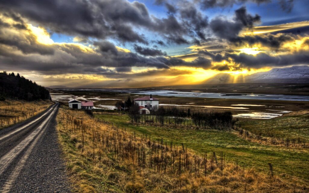 Gravel road at dawn winding through fields to cozy houses with mountains and glowing sun light Wallpaper