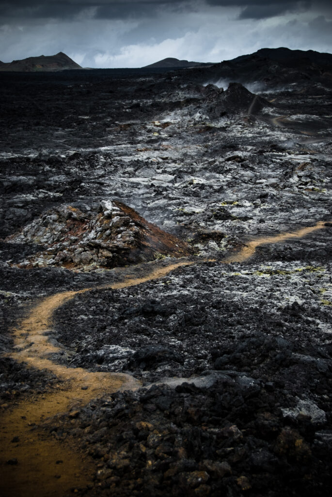 Winding golden earth path through dark volcanic rocks leading to shadowy mountains under moody sky Wallpaper