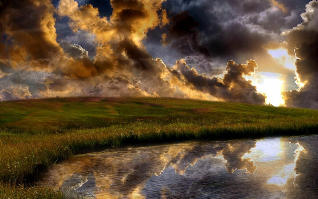 Dramatic evening clouds in gold and orange over grassy hill with calm water reflection wallpaper