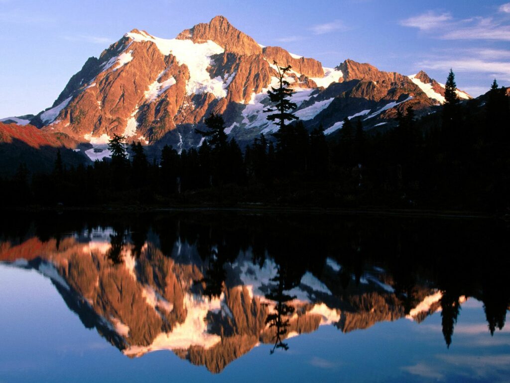Golden evening light on rugged mountains reflected in calm lake with dark foreground trees Wallpaper