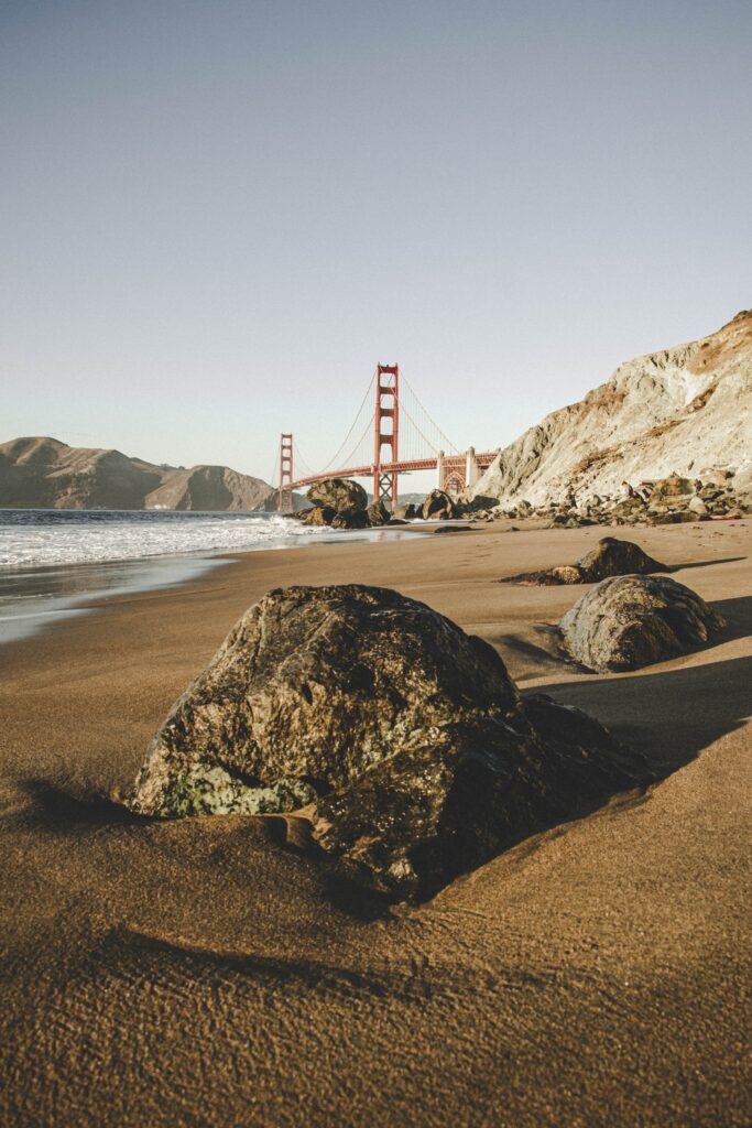 Golden Gate Bridge framed by coastal rocks on sandy beach under soft blue sky and sea background Wallpaper