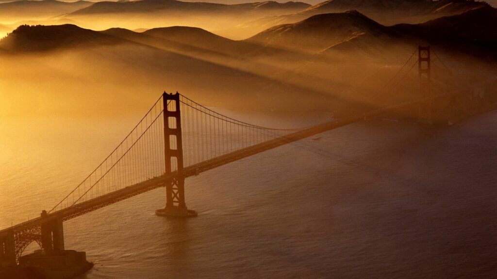 Aerial view of Golden Gate Bridge over calm waters with golden sunlight and misty hills background Wallpaper