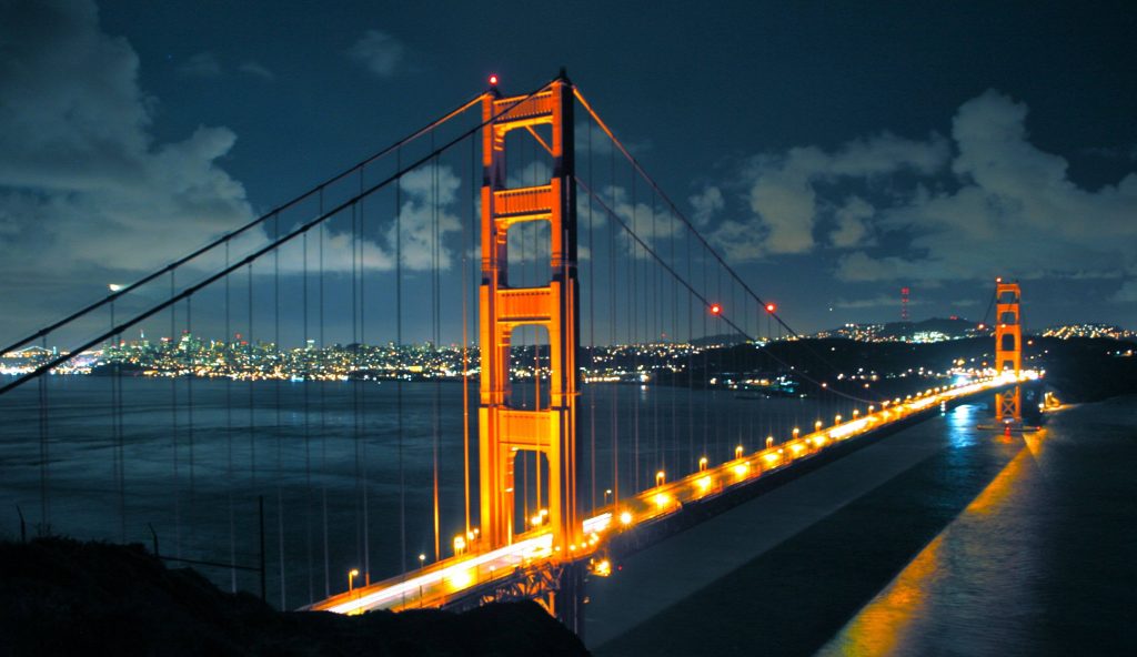 Golden Gate Bridge glowing orange at night with San Francisco skyline and cloudy blue sky Wallpaper