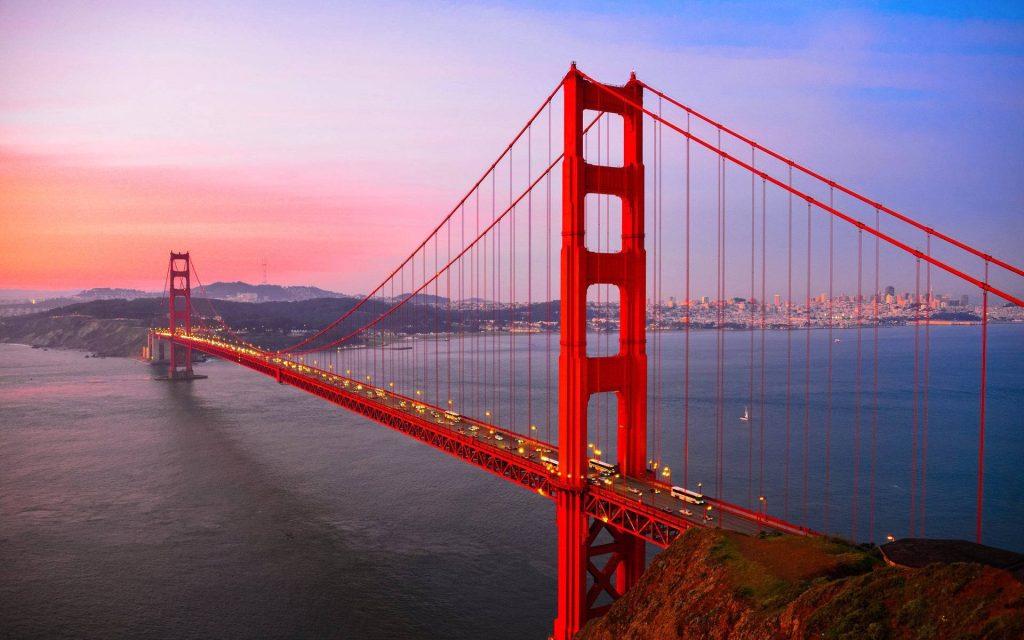 Golden Gate Bridge at sunset glowing in orange with San Francisco skyline and calm bay background Wallpaper