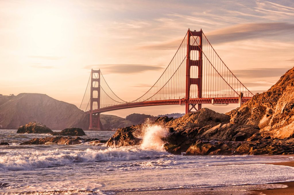 Golden Gate Bridge glowing in sunlight with waves crashing on rugged San Francisco shore wallpaper