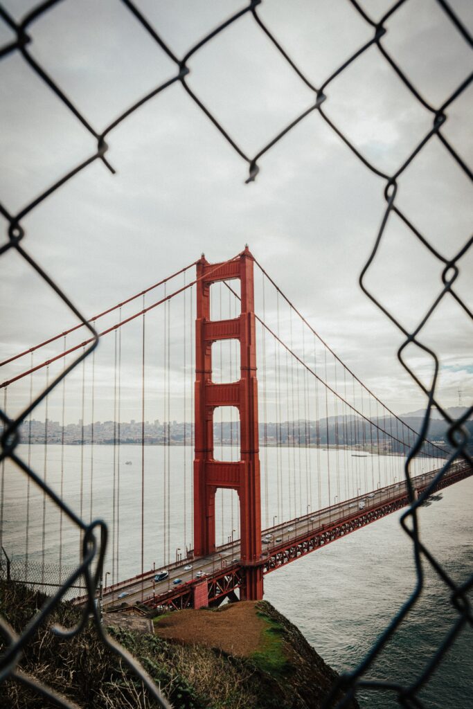 Red suspension bridge seen through broken mesh fence over calm sea and cloudy sky wallpaper