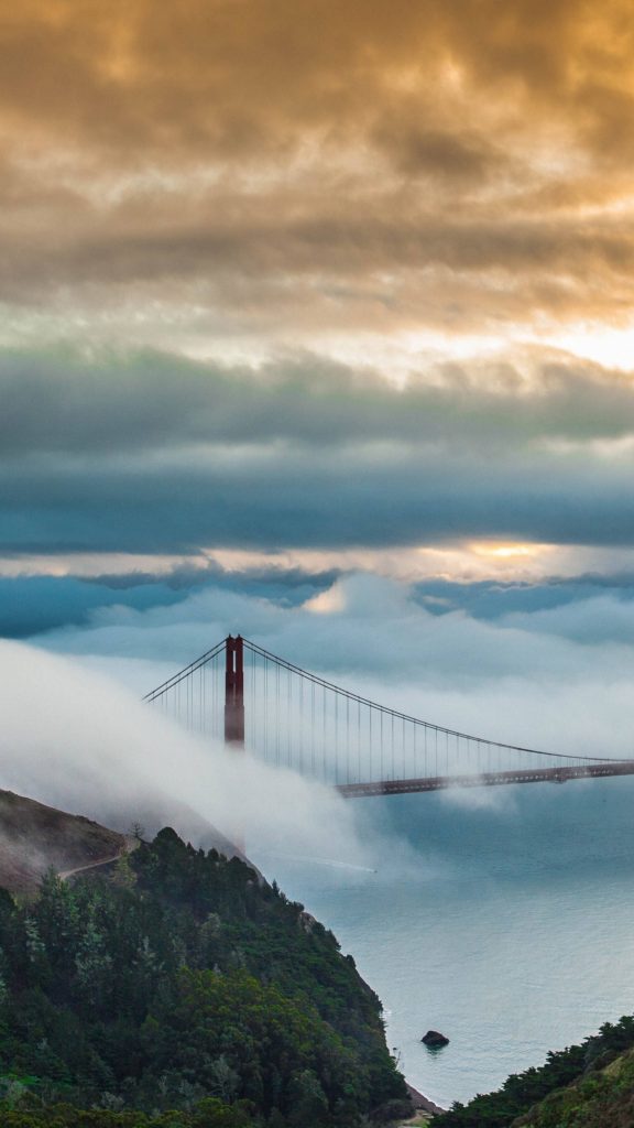 Golden Gate Bridge emerging from fog over calm waters with green cliffs and golden sunlight Wallpaper
