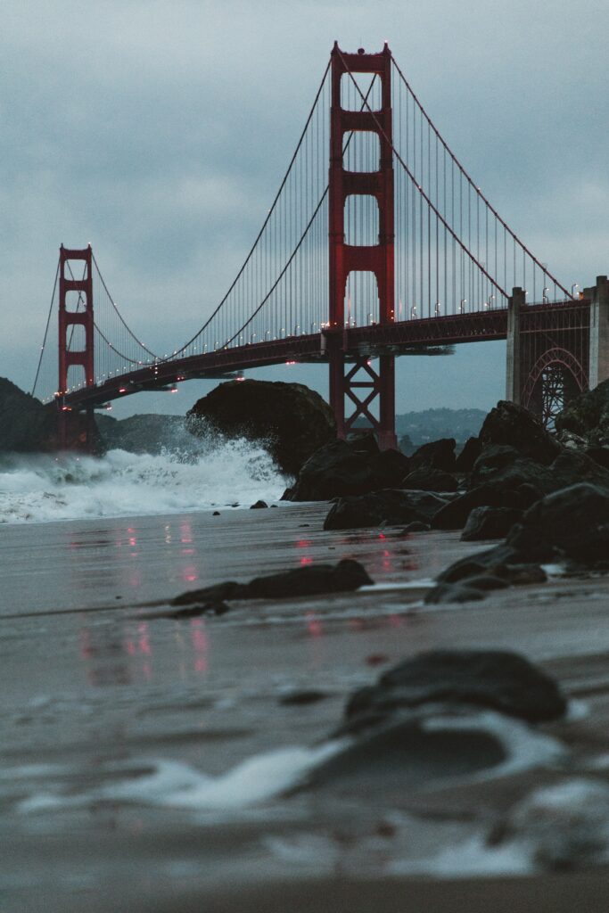 Golden Gate Bridge at dusk with red structure ocean foam on dark coastal rocks wallpaper