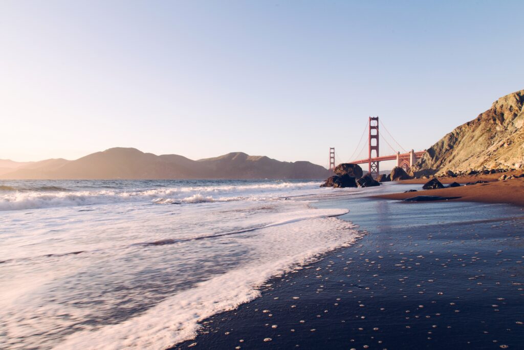 Golden sunlight over gentle sea waves with Golden Gate Bridge cliffs sand and blue sky wallpaper