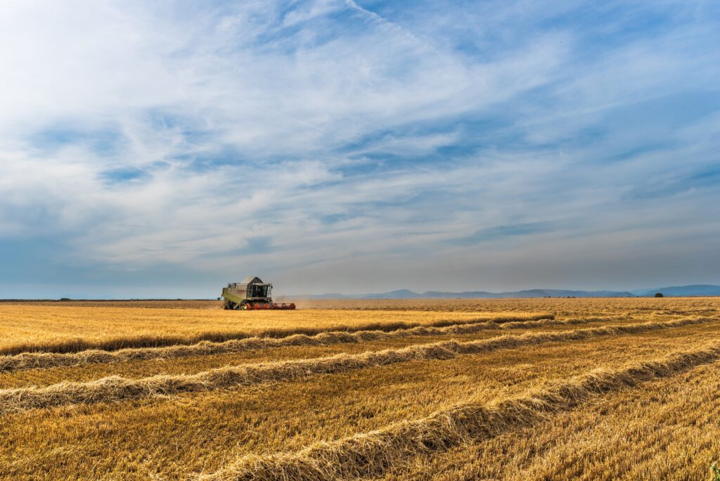 Lone harvester working in golden crop field under partly cloudy sky with distant hills background Wallpaper