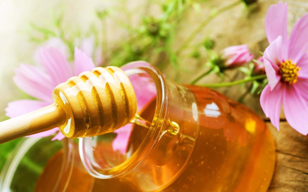 Closeup of golden honey dripping from wooden dipper into glass jar with pink Cosmos flowers Wallpaper