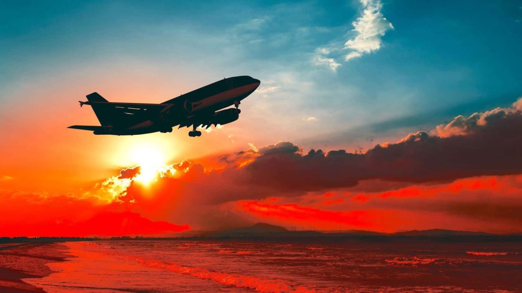 Plane silhouette flying over ocean at golden hour with fiery sunset sky and deep blue hues Wallpaper