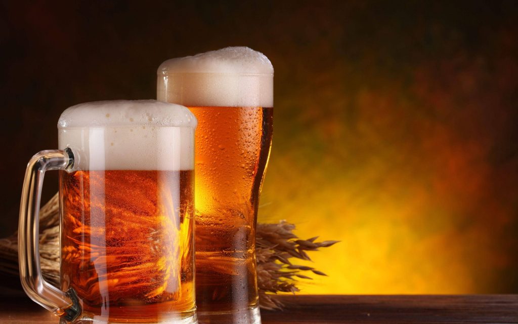 Two chilled golden lager glasses with foam and wheat stalks in warm ambient light background Wallpaper