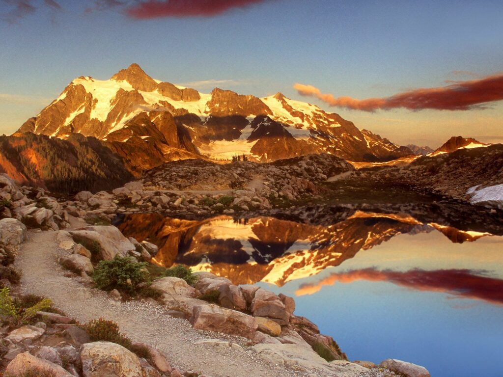 Snow-capped mountains at sunrise with rocky foreground and calm water reflecting vibrant sky Wallpaper