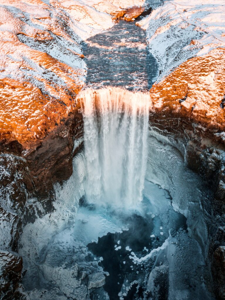 Icy waterfall flowing over snowy cliffs surrounded by frozen rocks illuminated by golden light Wallpaper