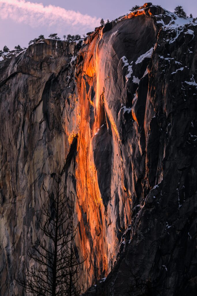Golden light illuminates rugged cliff with snow patches and trees in a dramatic mountain landscape Wallpaper