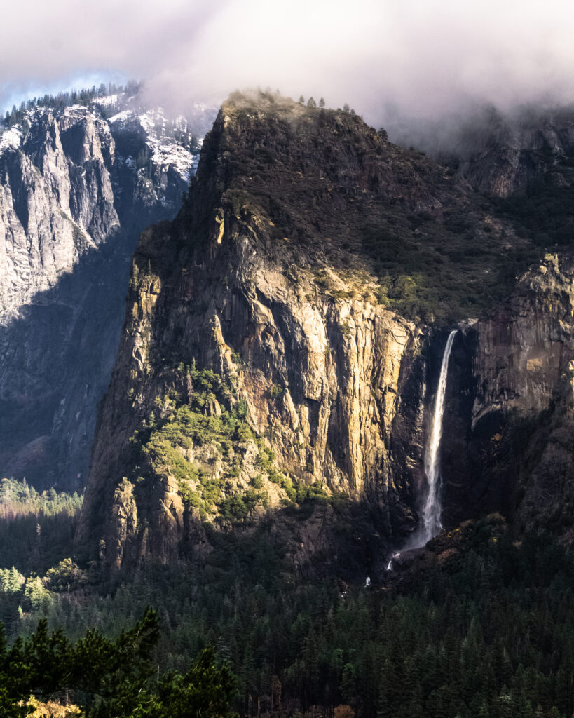 Towering mountain cliff with golden sunlight waterfall cascading into dense green forest misty peaks Wallpaper