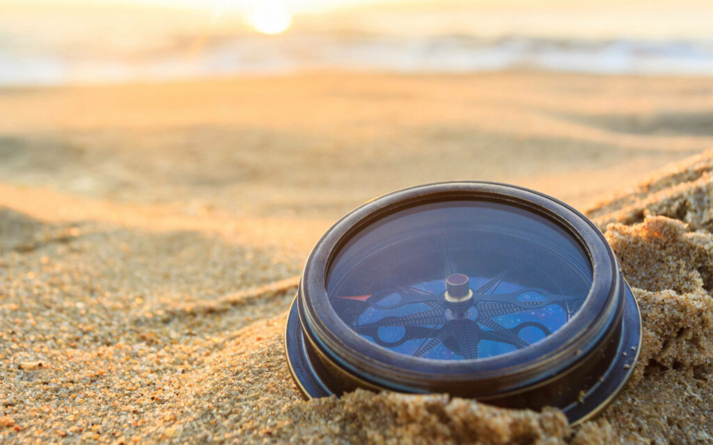 Classic compass in golden sand at sunset with blurred seascape for adventure and navigation Wallpaper