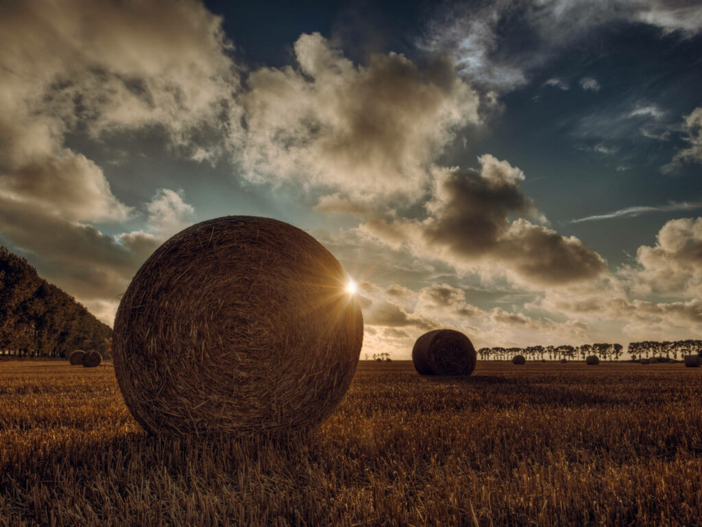 Golden straw bale in open field under dramatic sunset clouds rural agriculture wallpaper background