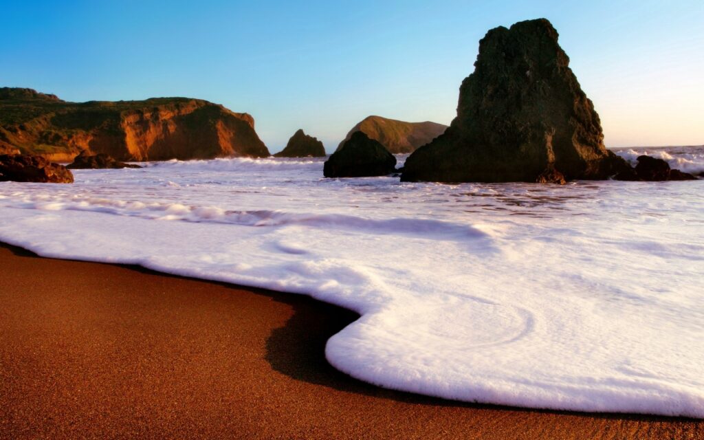 Golden sunlight on serene beach with foamy waves and large dark rocks at shoreline wallpaper