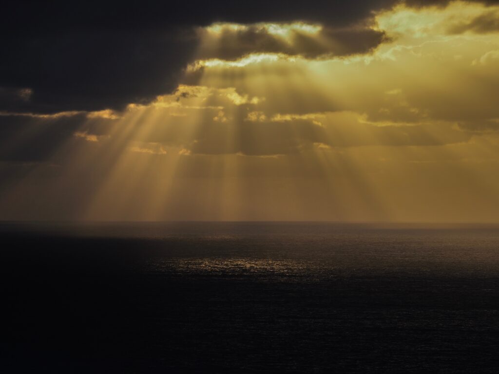 Golden sunlight piercing dramatic clouds over calm sea creating serene peaceful nature scene wallpaper