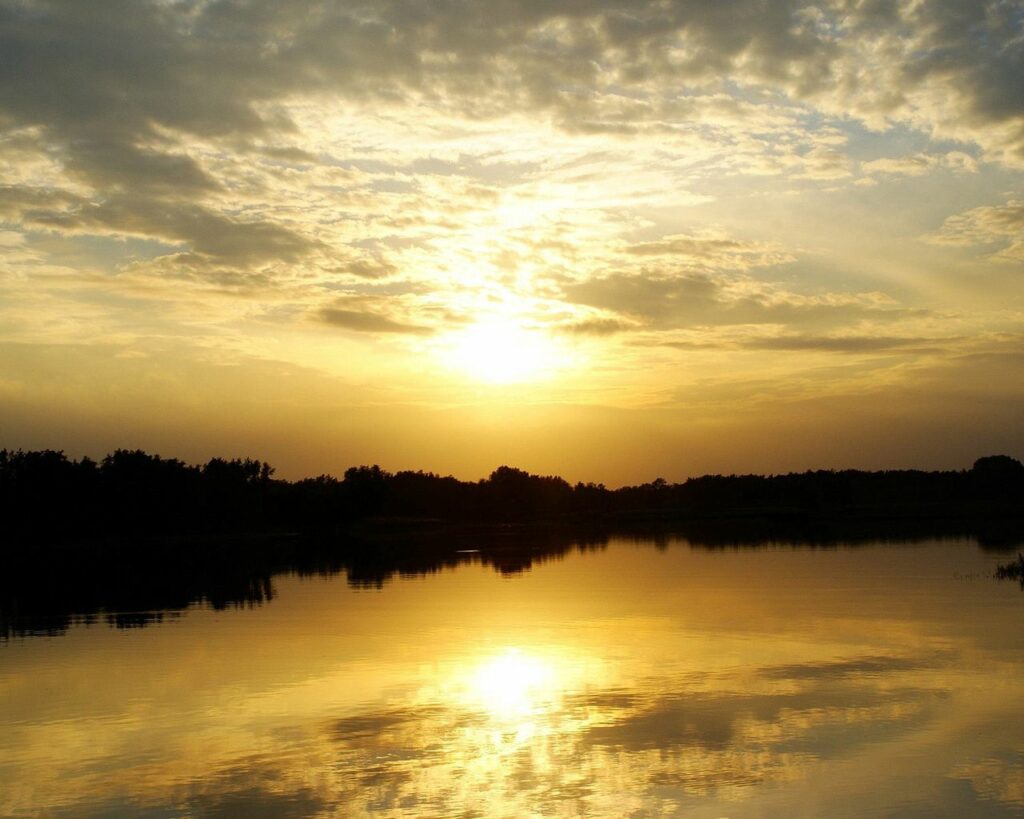 Sunlight filtering through clouds over calm lake reflecting trees serene natural wallpaper background