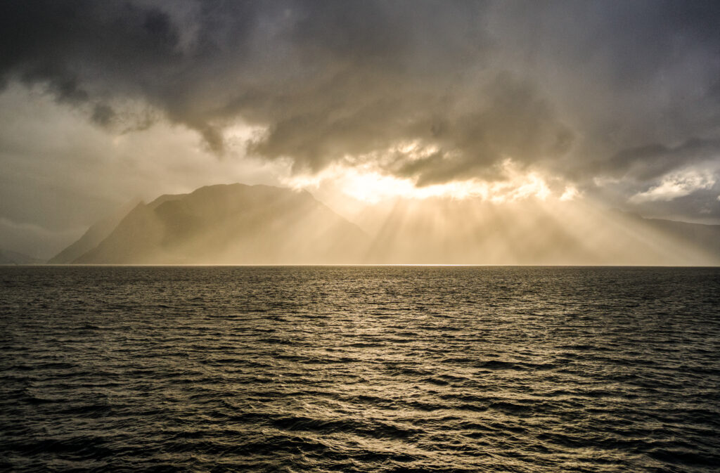 Golden sunlight pierces clouds over rippling sea with distant mountains serene nature view Wallpaper