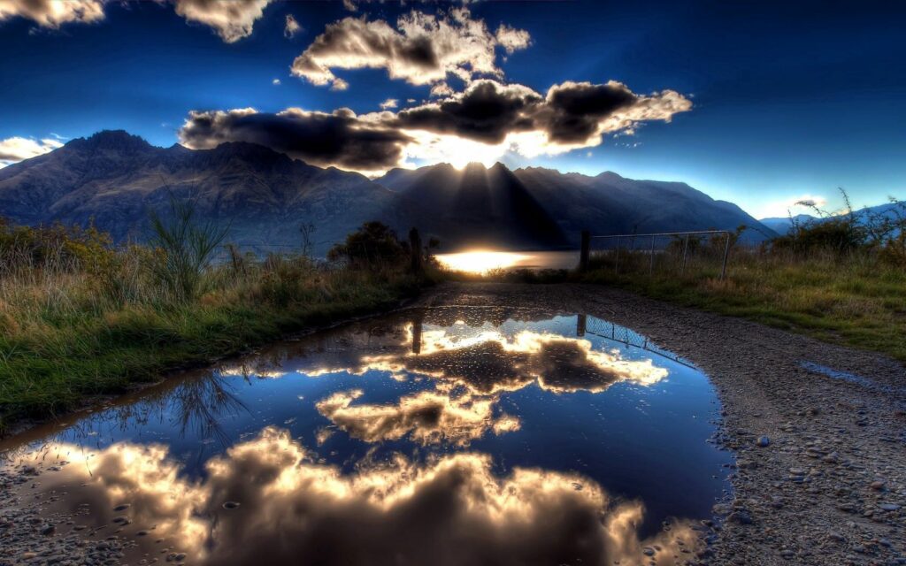 Sunlight through dramatic clouds over mountains with golden lake reflections and vibrant blue sky Wallpaper