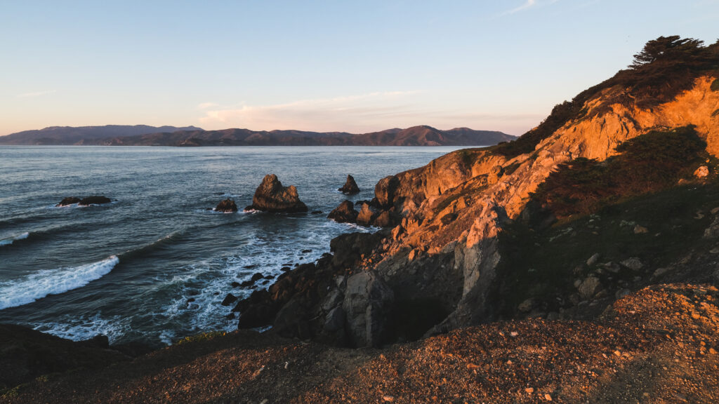 Golden sunlight on rugged coastal cliffs and rocks overlooking serene blue sea with gentle waves Wallpaper
