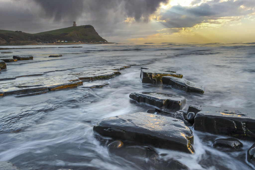 Golden sunlight over rugged coastline with smooth dark stones and swirling shallow sea shoreline Wallpaper