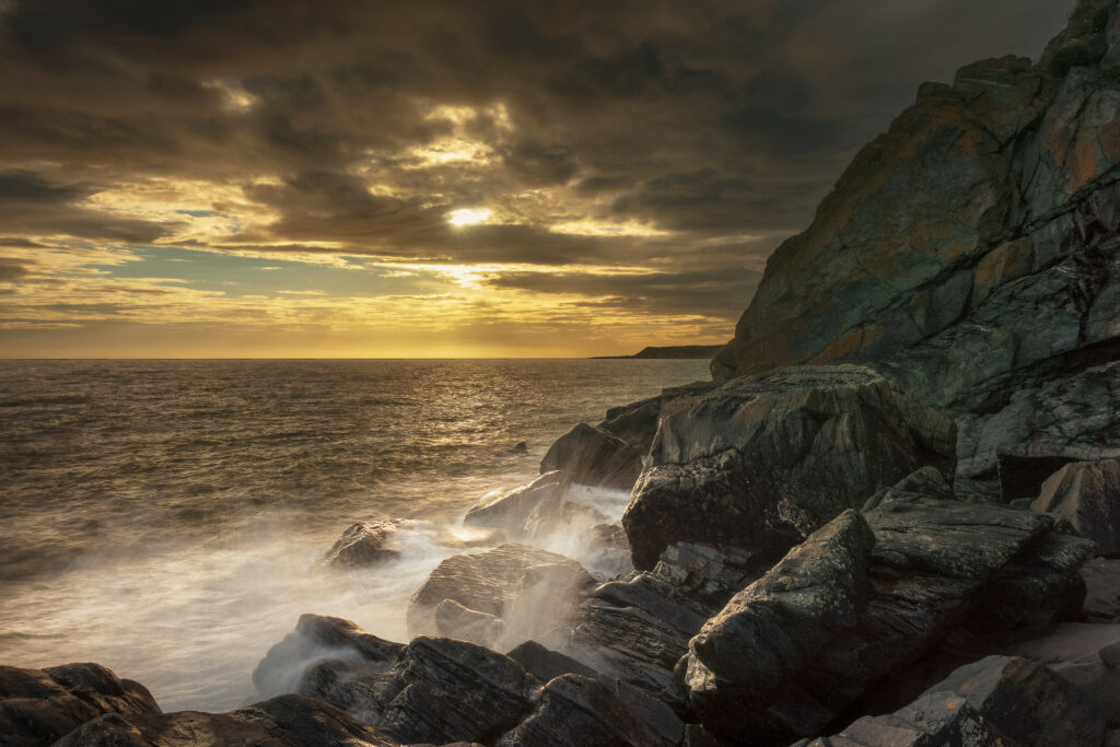 Golden sunlight illuminates rugged coastal rocks and dynamic ocean splashes under dramatic clouds Wallpaper