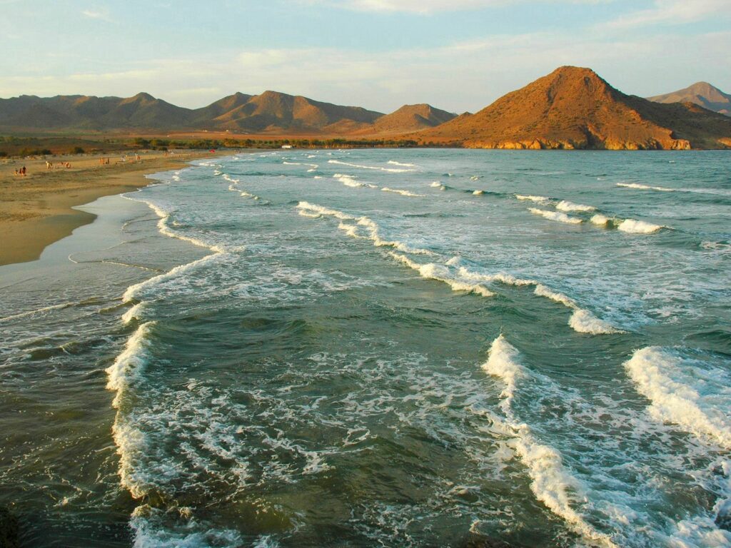 Golden sunlight on gentle waves at a serene Spanish beach with rugged mountains backdrop Wallpaper