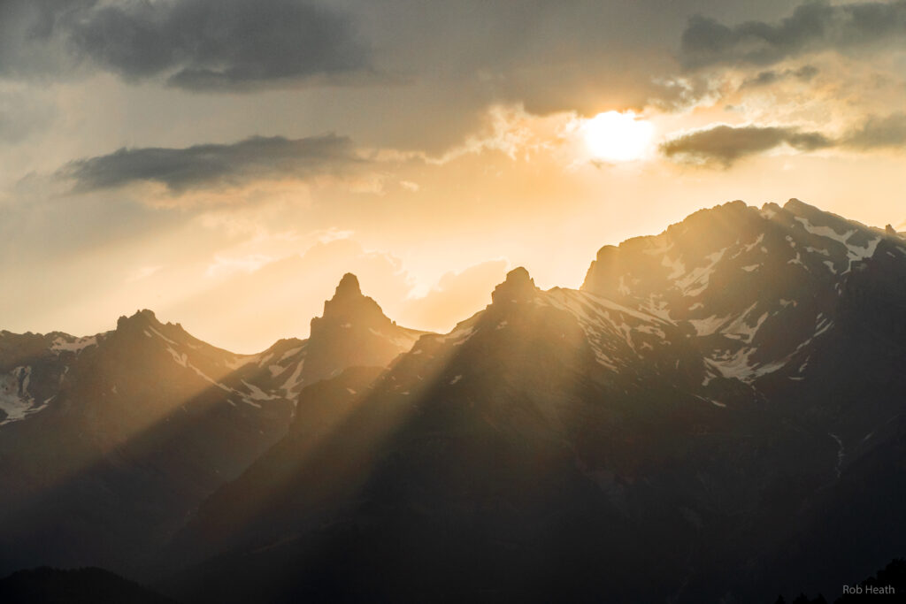 Golden sunlight rays shine through clouds over rugged snow-capped mountain peaks landscape Wallpaper