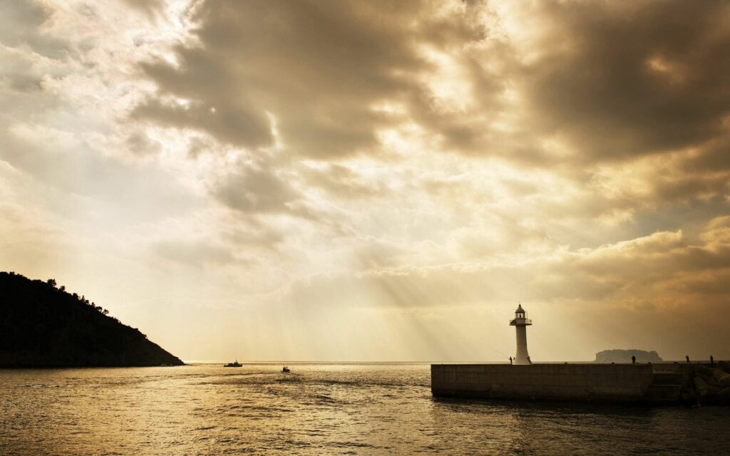 Golden sunlight through clouds brightens lighthouse on stone pier over calm sea with island boats Wallpaper