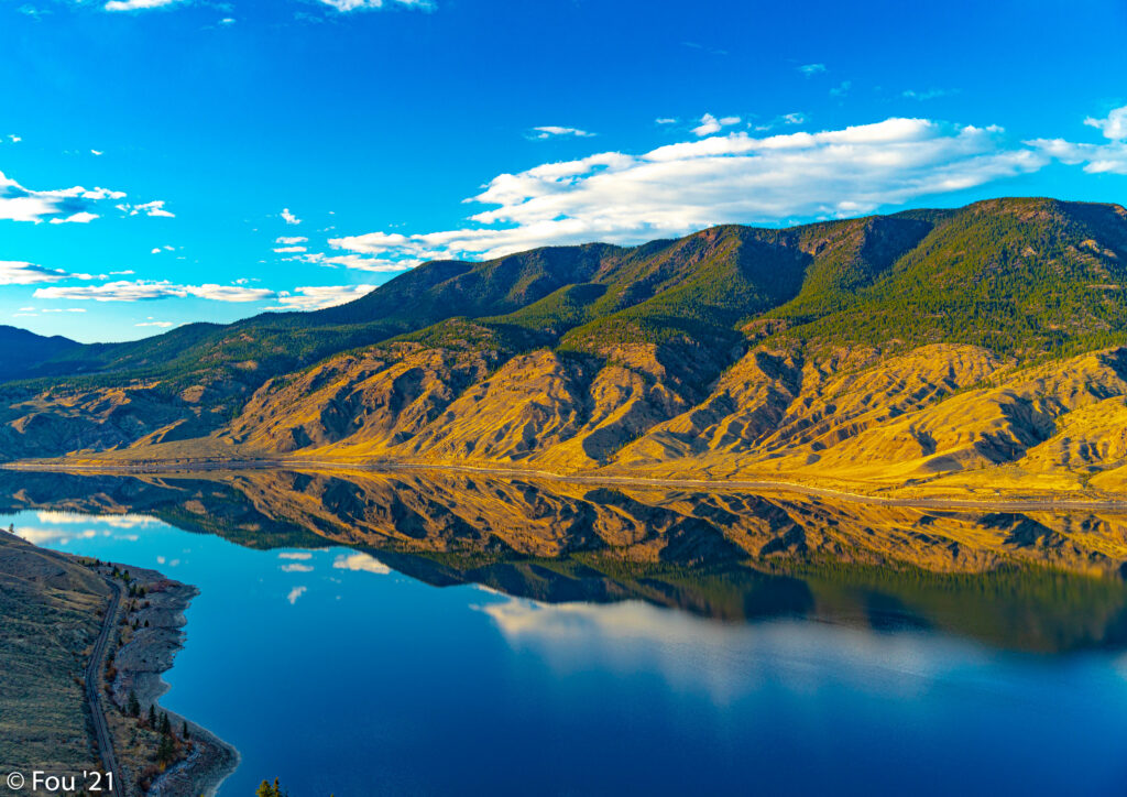 Rugged mountains in golden sunlight reflected in calm river under vibrant blue sky with clouds Wallpaper
