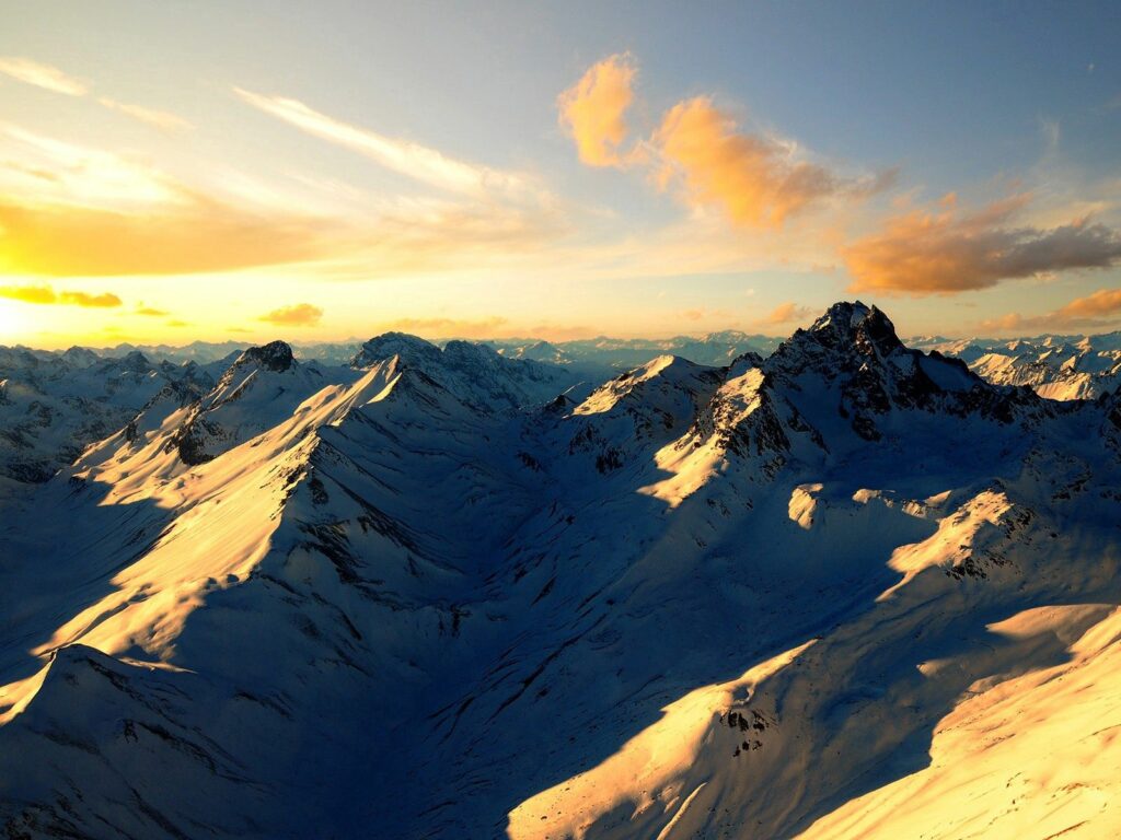 Snowy mountain peaks bathed in warm golden sunlight with dramatic shadows and vibrant sky Wallpaper