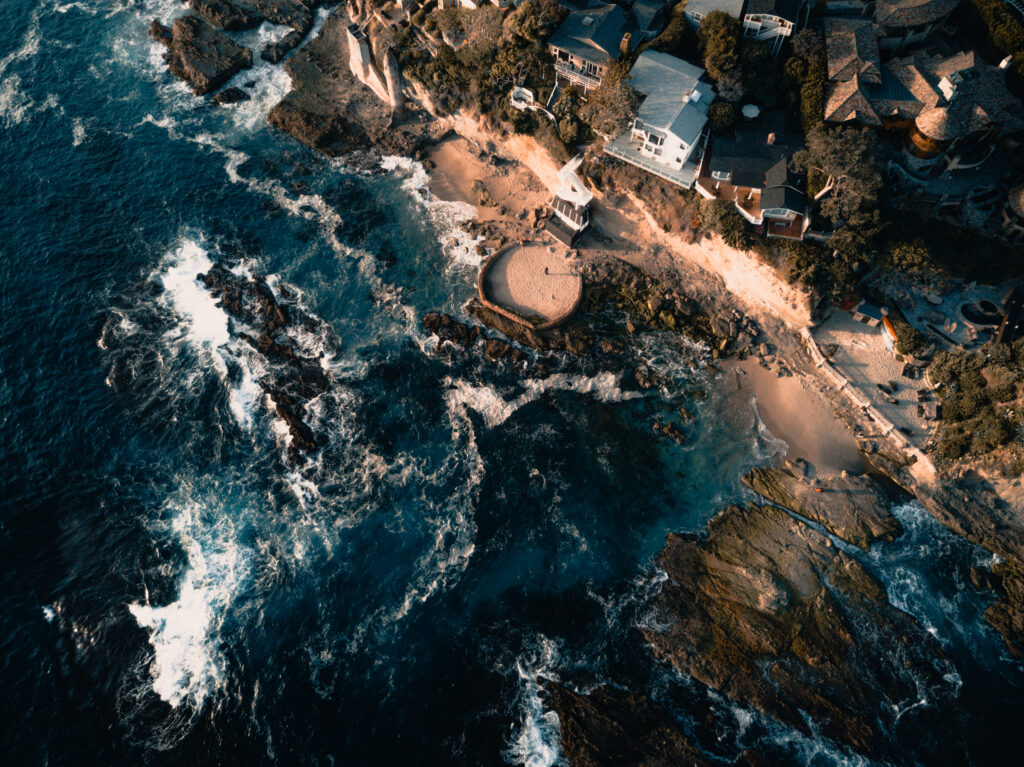 Aerial view of sea waves crashing on rocky shore with sandy beaches and coastal buildings sunlight Wallpaper