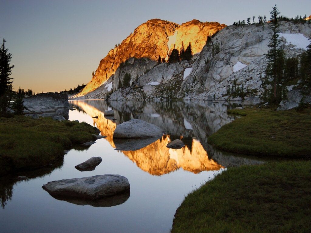 Serene mountain cliffs in golden sunlight reflected in calm lake with green grass and pine trees Wallpaper