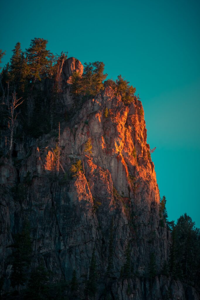 Rugged mountain peak in golden sunlight with textured rocks and scattered trees under blue sky Wallpaper
